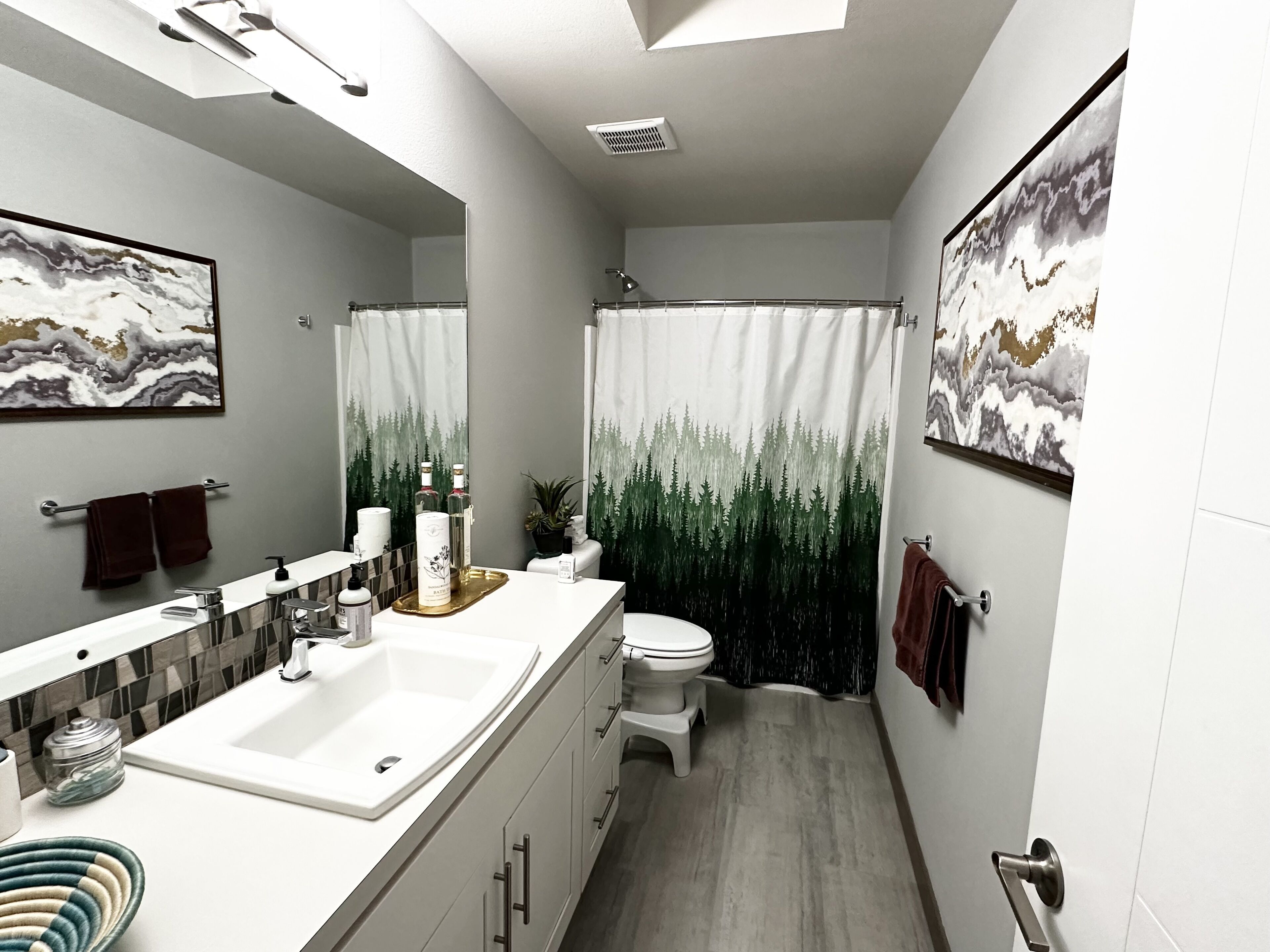 A modern bathroom with a large mirror, a white sink, a toilet, and a shower curtain with a forest design.