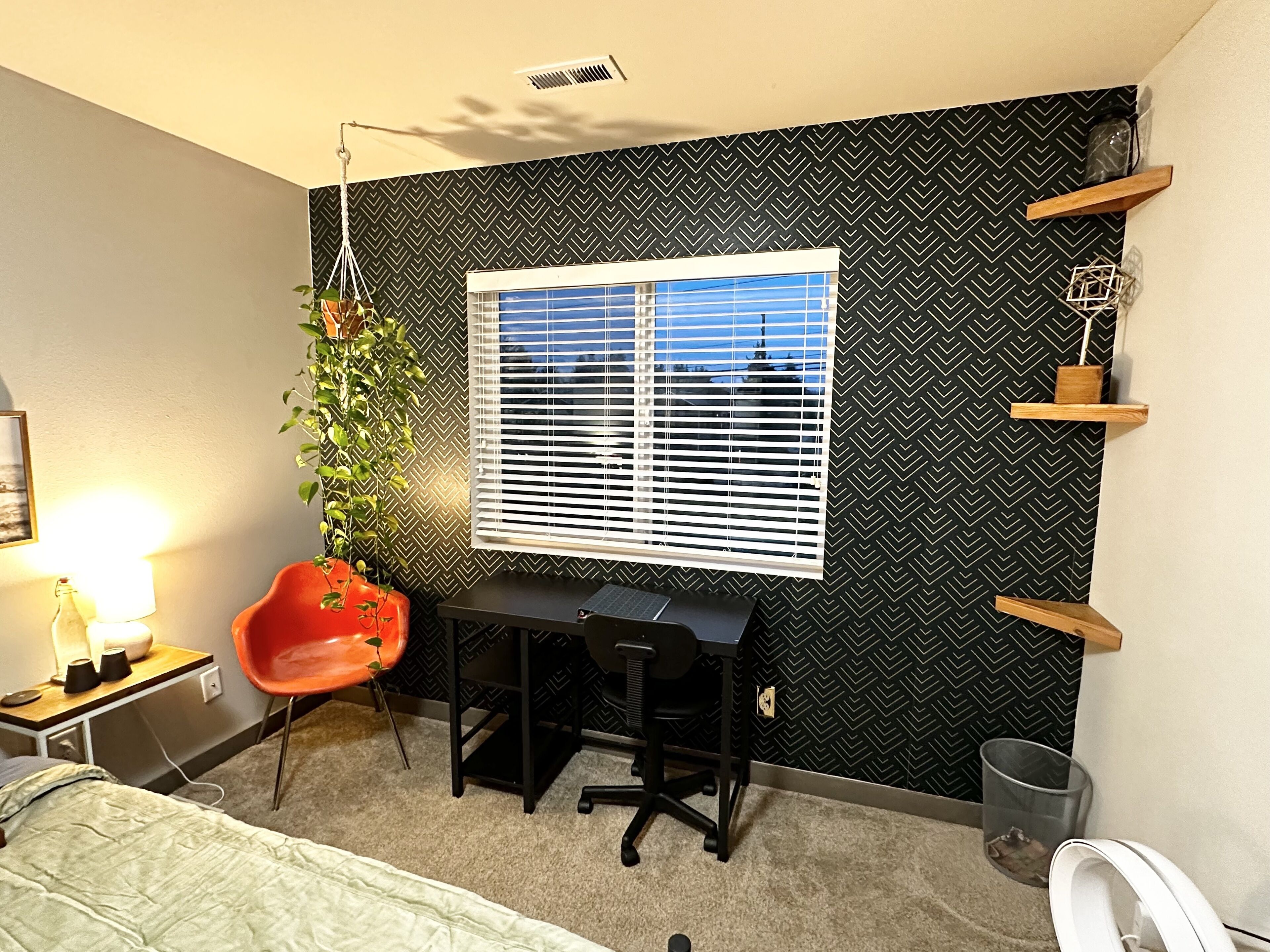 Ein gemütliches Schlafzimmer mit Schreibtisch, einer hängenden Pflanze, einem Sessel, einem Bett und einem Fenster mit Jalousien.