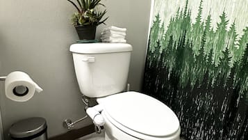 A bathroom with a toilet, a trash bin, a toilet paper holder, a small potted plant, and a shower curtain with a forest design.
