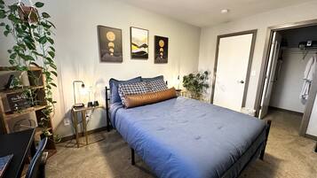 A bedroom with a bed, a desk, a chair, a hanging plant, and a wall with three framed pictures.