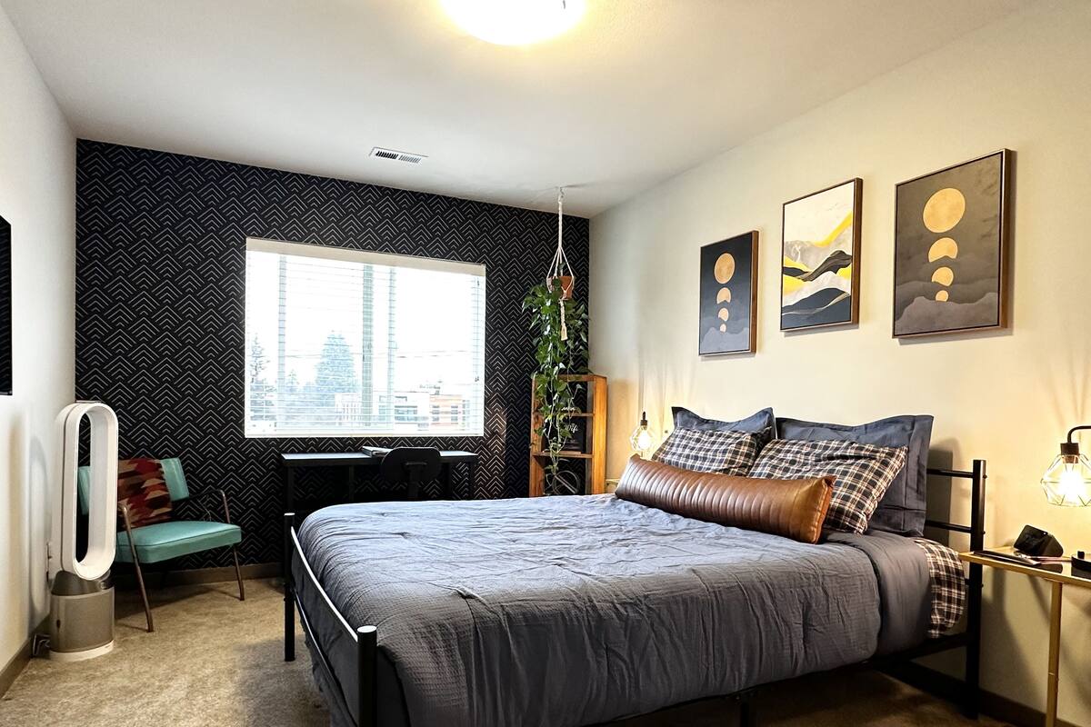 Ein modernes Schlafzimmer mit einem großen Bett, einem Schreibtisch und einem Stuhl. Ein Fenster bietet Blick auf die Stadt, und die Wand ist mit einem schwarz-weißen Muster versehen.