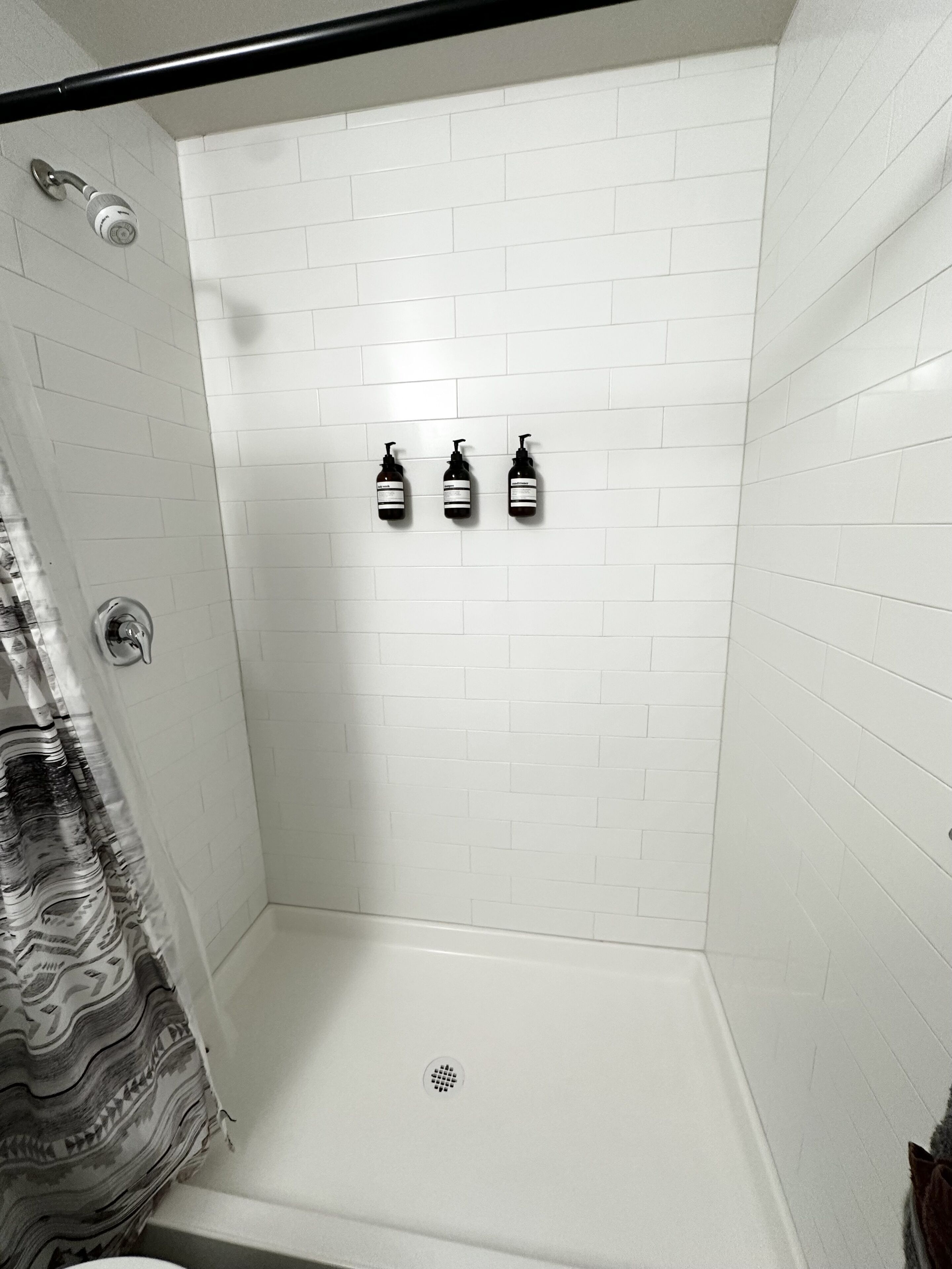 A shower with white tiled walls, a shower curtain, and three bottles of shampoo.