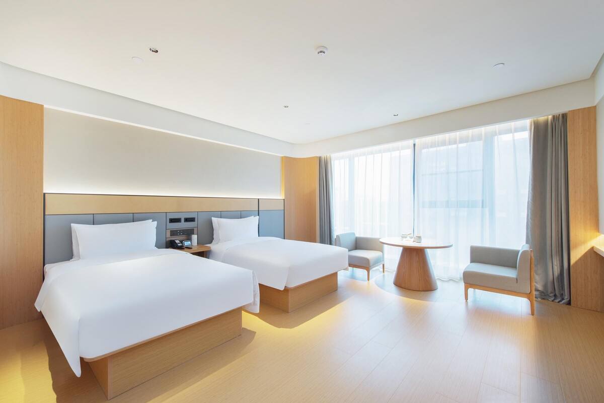 A hotel room with two beds, a small table, a chair, and a large window with curtains.