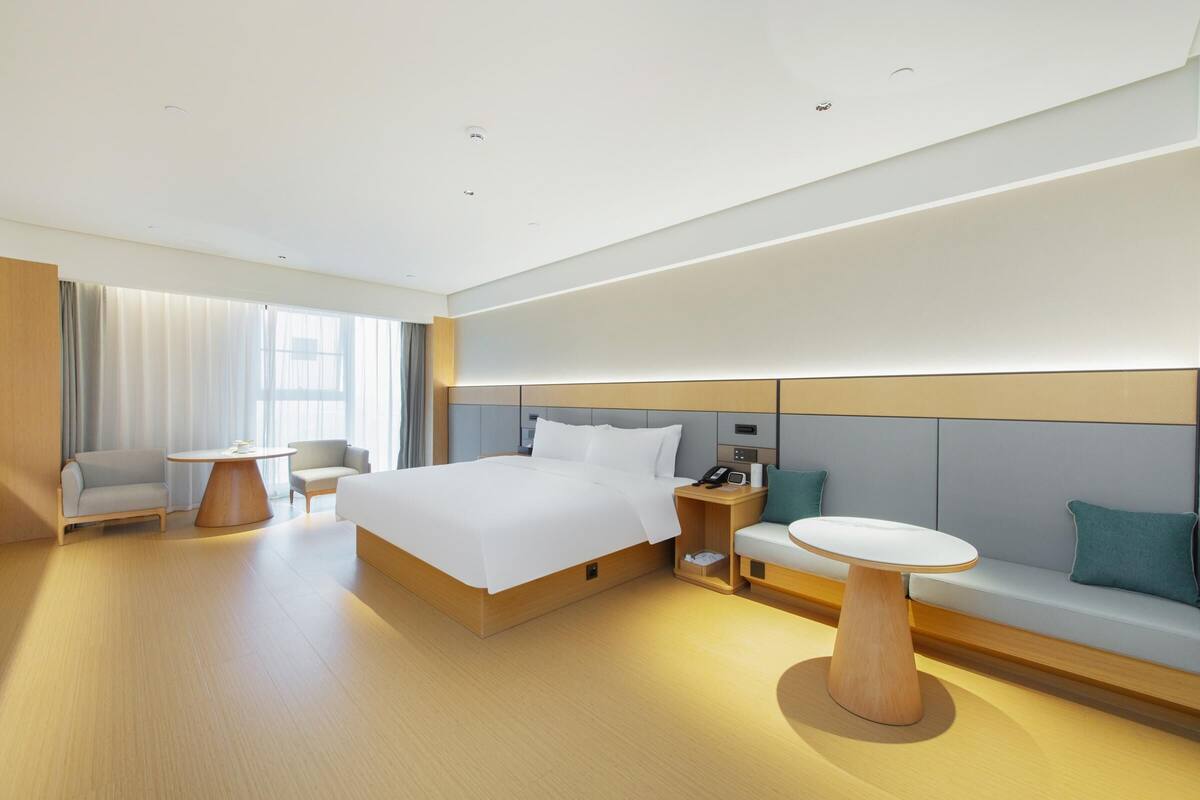 A modern hotel room with a large bed, a bedside table, a chair, a sofa, and a small table.