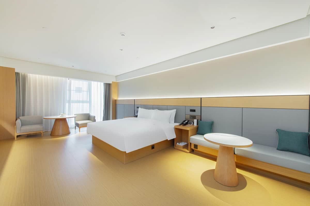 A modern hotel room with a large bed, a bedside table, a chair, a sofa, and a small table.