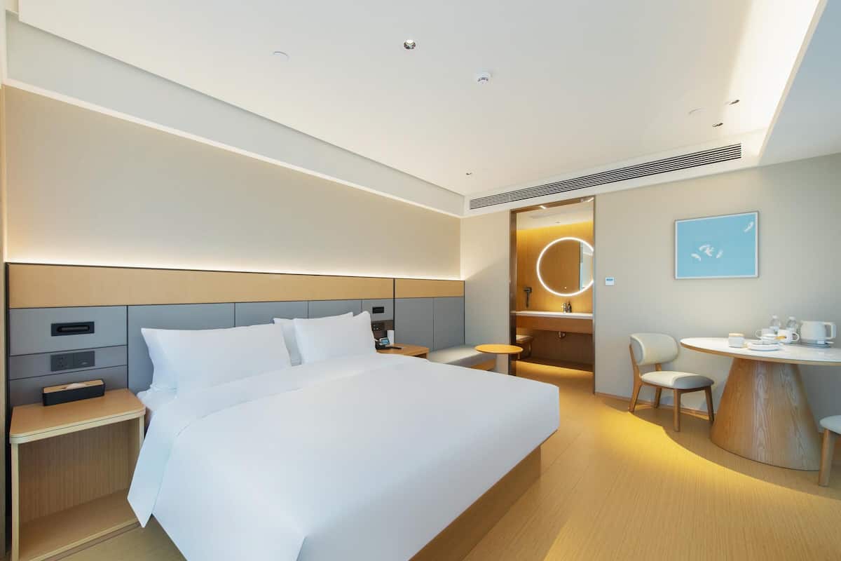 A modern hotel room with a large bed, bedside tables, a desk with a chair, and a small round table with a chair.