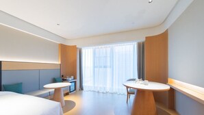Habitación de hotel con cama, dos mesas redondas, un escritorio pequeño y un ventanal con cortinas traslúcidas.