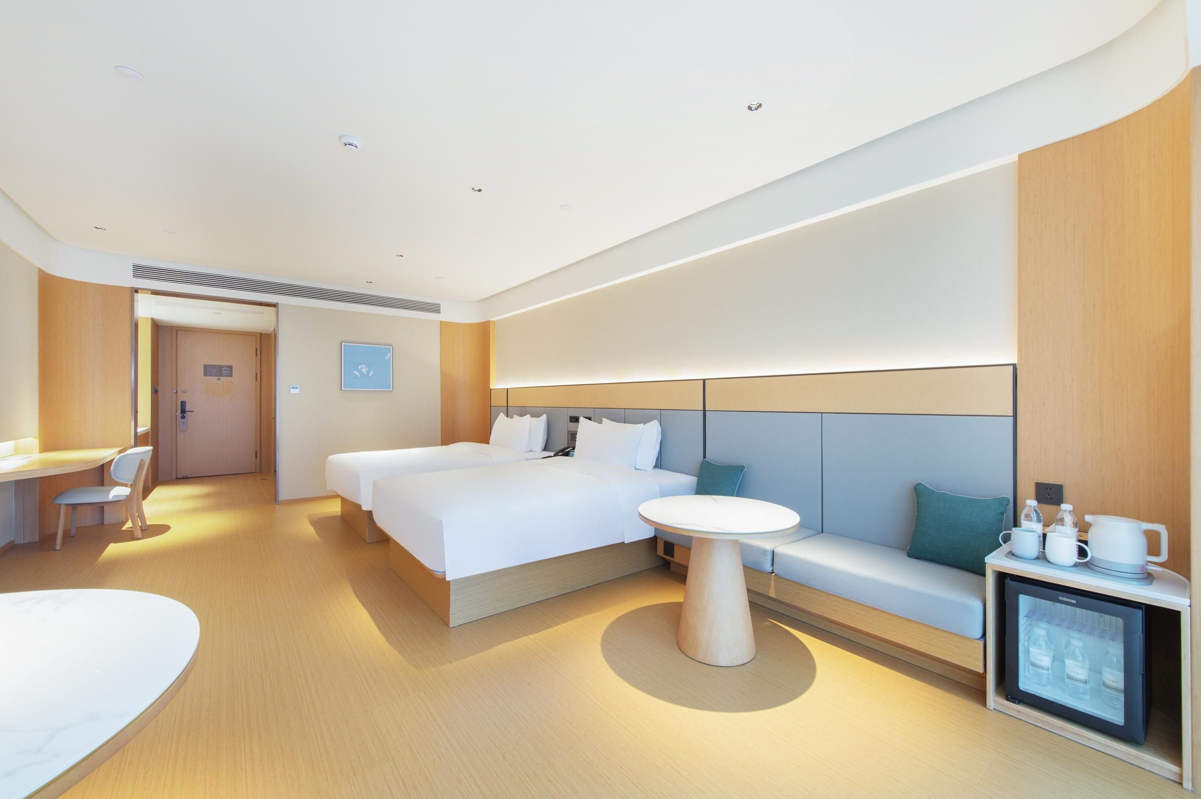 A modern hotel room with a large bed, a small round table, a sofa, a desk, and a mini-fridge.