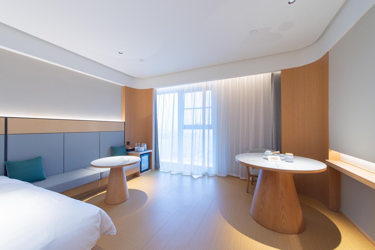 A modern hotel room with a bed, two round tables, a sofa, a desk, and a large window with sheer curtains.