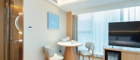 A modern hotel room with a round dining table, two chairs, a flat-screen TV, and a minibar.