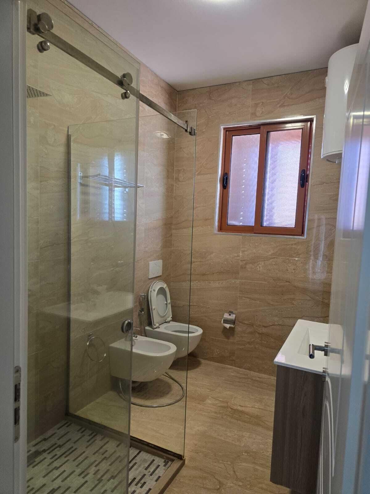 Un baño moderno con ducha envuelta en vidrio, dos inodoros, un lavamanos y una ventana.