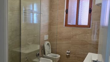 Un baño moderno con ducha envuelta en vidrio, dos inodoros, un lavamanos y una ventana.