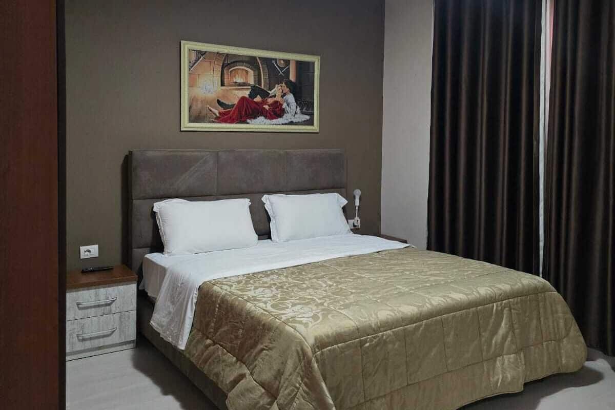 Un dormitorio con cama, mesita de noche, un cuadro en la pared y una ventana con cortinas.