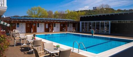 A swimming pool with lounge chairs and tables, a building with a sloped roof, and a clear blue sky.
