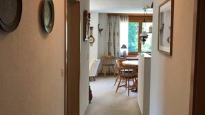 Ein Flur, der in einen Essbereich mit Tisch und Stühlen führt. An der Wand hängt eine Uhr, und ein Fenster ist mit Vorhängen versehen.