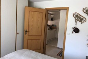 A bedroom with a wooden door, a bed, and a small kitchen area visible through the door.