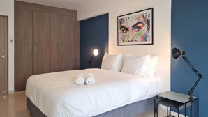 A neatly made bed with white linens and two towels, a bedside table with a lamp, a large wooden wardrobe, and a colorful abstract painting on the wall. - w* | Timeless 2BR In La Cresta (Panamá)