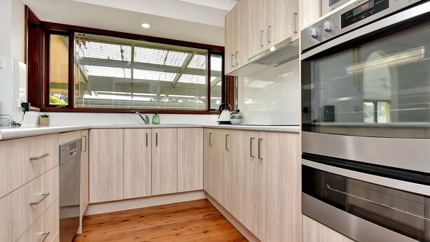 A modern kitchen with wooden cabinets, a stainless steel oven, and a window with blinds. - Breezy Bayside Haven at Brightwaters (Brightwaters)