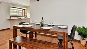 A dining area with a wooden table set for four, a kitchen in the background, and a potted plant on the floor. - Breezy Bayside Haven at Brightwaters (Brightwaters)