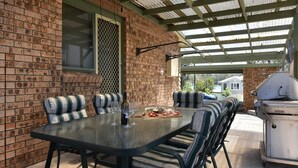 A covered patio with a brick wall, a table with chairs, a grill, and a window with a mesh screen. - Breezy Bayside Haven at Brightwaters (Brightwaters)