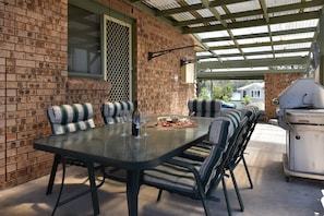 Een overdekt terras met een bakstenen muur, een tafel met stoelen, een barbecue en een raam met hor.
