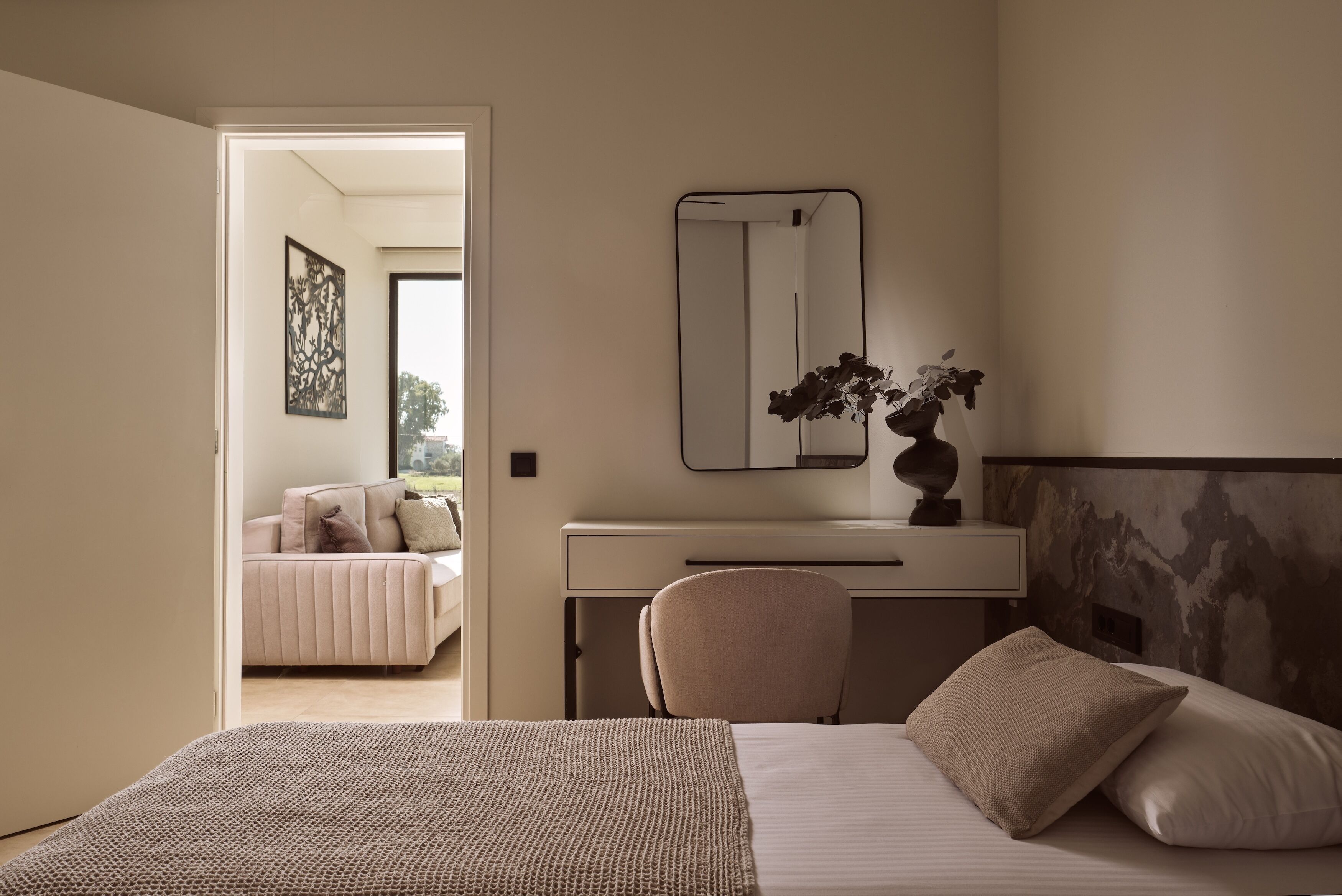 A bedroom with a bed, a desk, a chair, a mirror, and a view of another room.