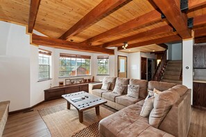 A spacious living room with a high ceiling, wooden beams, a large sofa set, a coffee table, and a view of the outdoors through large windows.