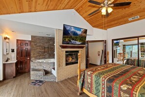 A room with a fireplace, a bed with a patterned bedspread, a television mounted above the fireplace, and a ceiling fan.