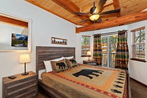 Une chambre avec un plafond en bois, un lit recouvert d’une couette à motifs d’ours, une table de chevet avec une lampe et une fenêtre avec des rideaux.