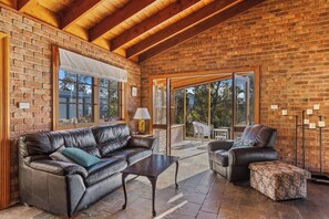 A spacious living area with a brick wall, leather sofas, a coffee table, and a view through large glass doors to an outdoor patio. - Country style Stunner Bushland Peace and Privacy (Margate)