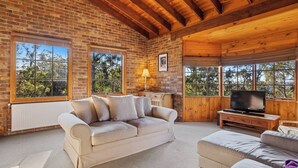 Una habitación acogedora con pared de ladrillo, vigas de madera en el techo, un sofá color beige, un televisor y ventanas amplias con vistas a árboles.