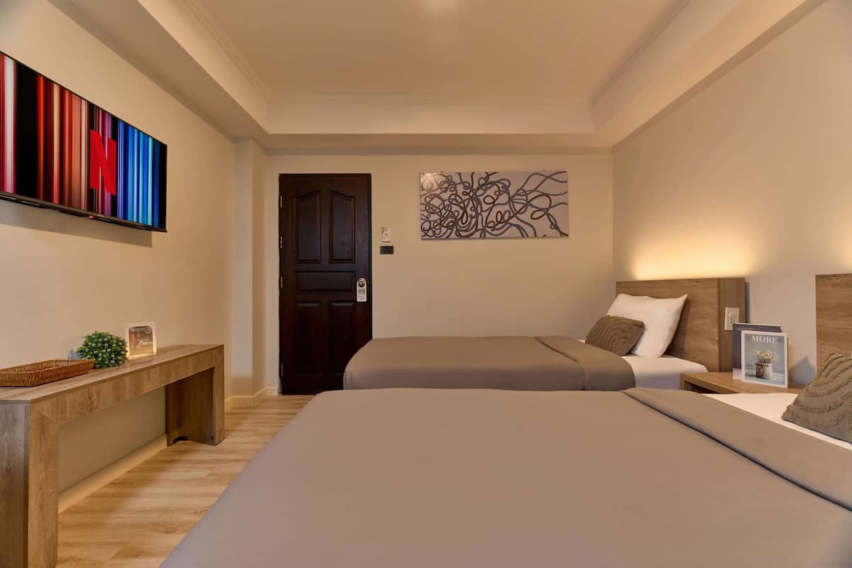 Habitación de hotel con dos camas, un banco de madera, un televisor con un logotipo colorido, arte abstracto en la pared y una puerta con marco oscuro.