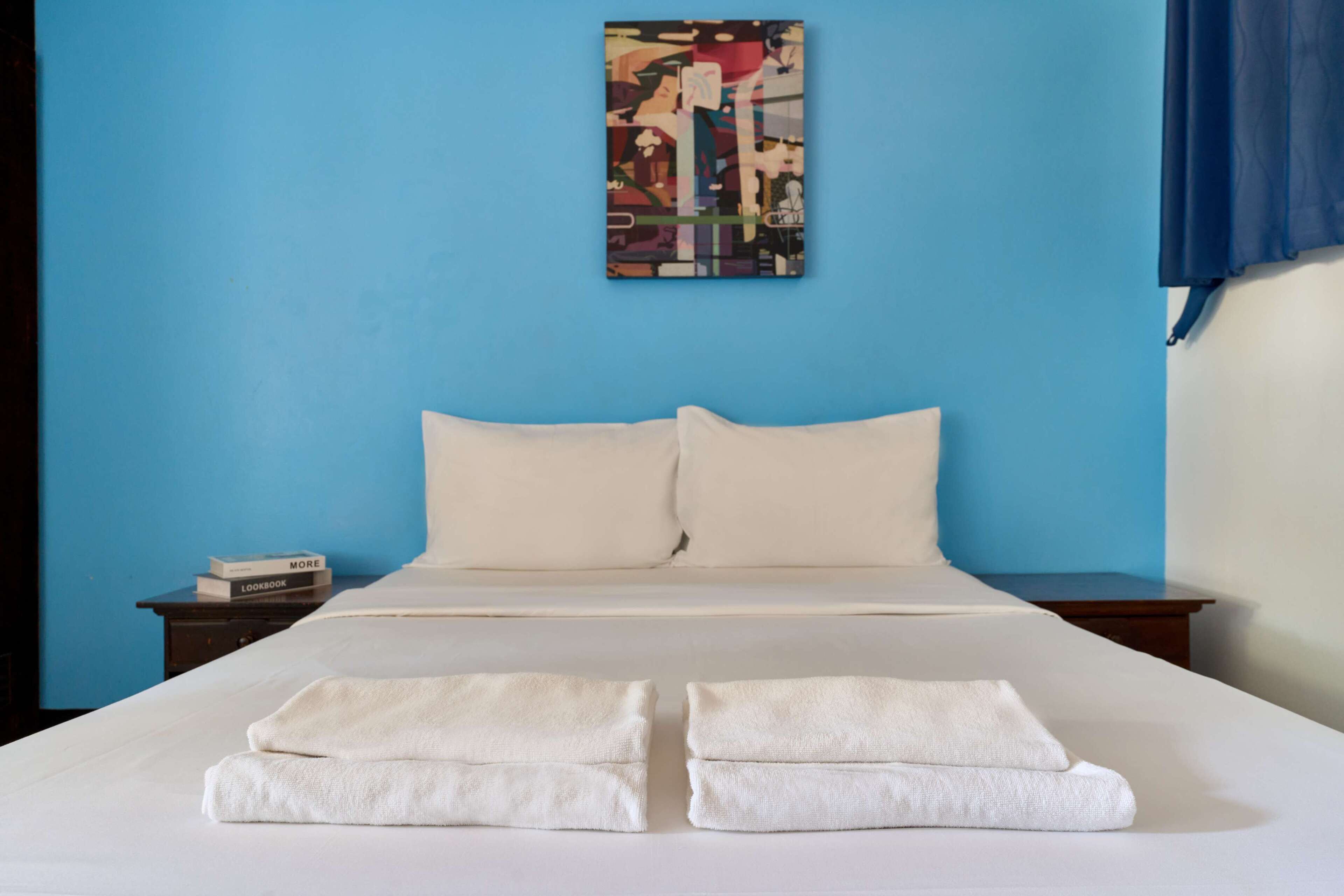 A neatly made bed with white linens and towels, a blue wall, and a small bedside table with a book.
