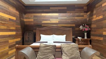 Un dormitorio con pared de listones de madera, estructura de cama de madera, mesita de noche y mesita auxiliar con un jarrón de flores, además de un tragaluz sobre la cama.