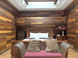 Un dormitorio con pared de listones de madera, estructura de cama de madera, mesita de noche y mesita auxiliar con un jarrón de flores, además de un tragaluz sobre la cama.