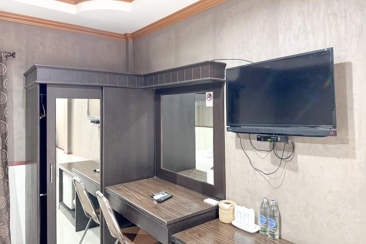 A hotel room with a large mirror, a flat-screen TV mounted on the wall, a wooden desk, a chair, a telephone, and two bottles of water.