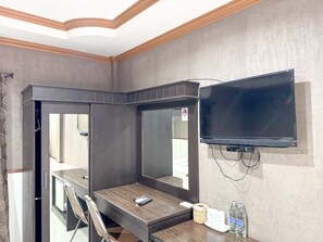 A hotel room with a large mirror, a flat-screen TV mounted on the wall, a wooden desk, a chair, a telephone, and two bottles of water. - Grand Amazon Hotel by Zuzu (Lampang)