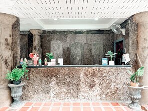 Mostrador de recepción de piedra con un ventilador, plantas en macetas y una puerta a una habitación.