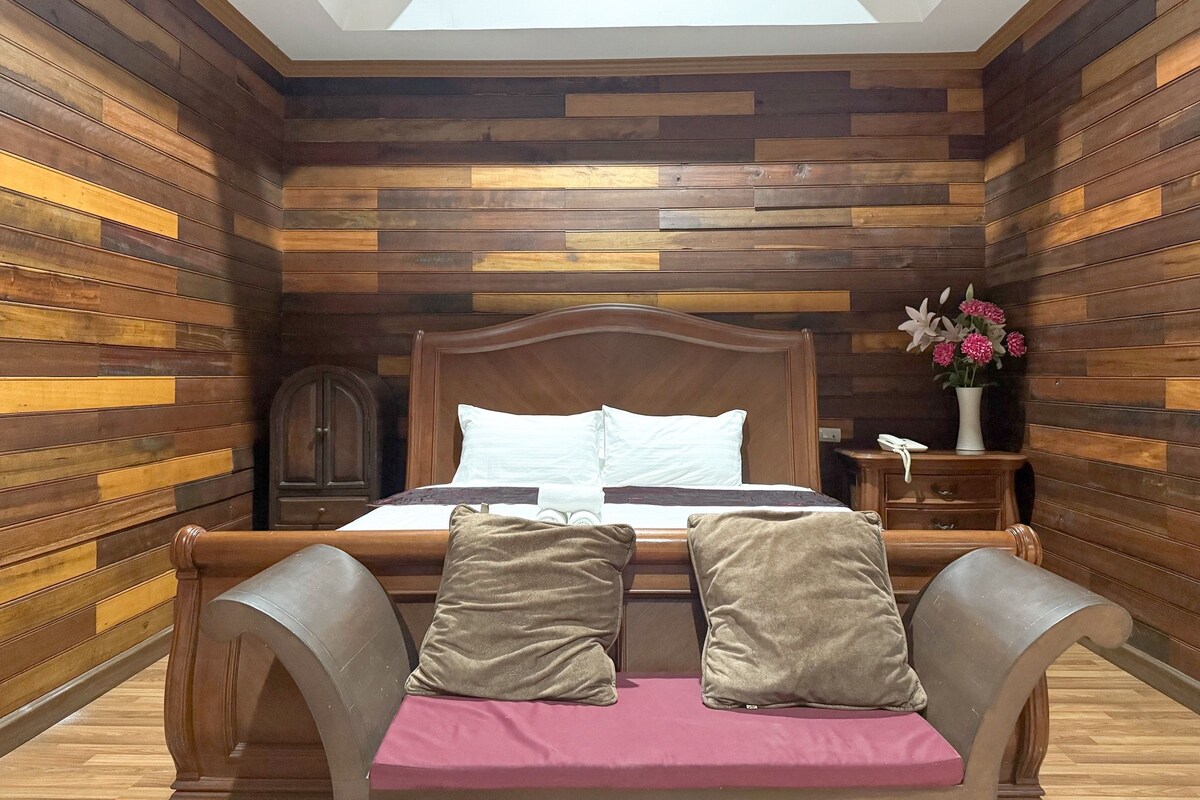 A bedroom with a wooden slat wall, a wooden bed frame, a nightstand, a bedside table with a vase of flowers, and a skylight above the bed.