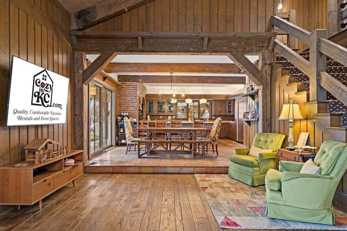 A rustic interior with a wooden dining area, a staircase, and a seating area with green armchairs.