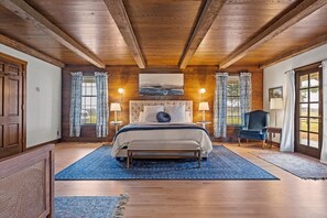Un dormitorio con una cama grande, vigas de madera en el techo, dos lámparas de noche y vistas al exterior por una ventana.