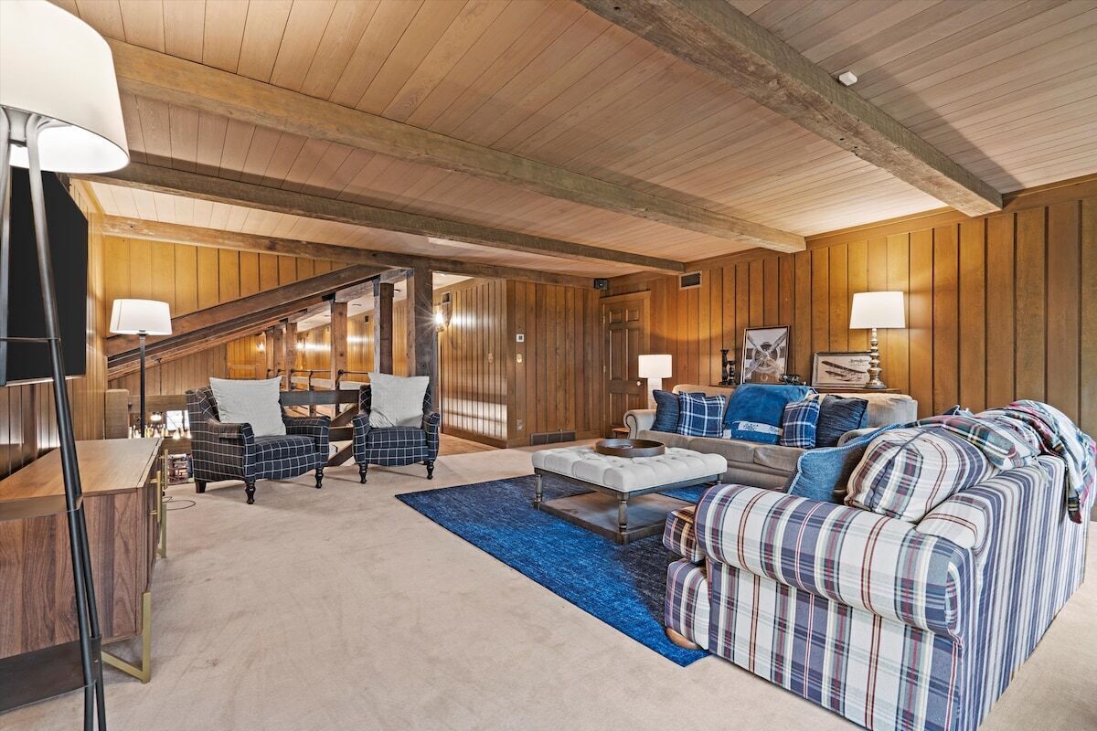 A spacious living room with a high ceiling, wooden paneling, a staircase, and a mix of seating including a sofa and armchairs.