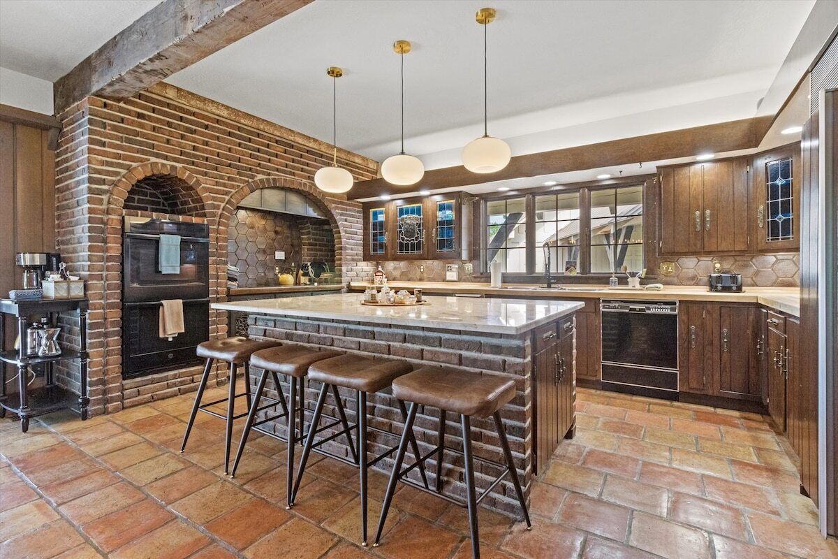 A kitchen with a brick wall, a central island with stools, hanging pendant lights, and a built-in oven.