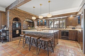 A kitchen with a brick wall, a central island with stools, hanging pendant lights, and a built-in oven. - Landing(43M0)- Sleeps 20 + Airway Strip + Hot Tub (Pleasant Hill)