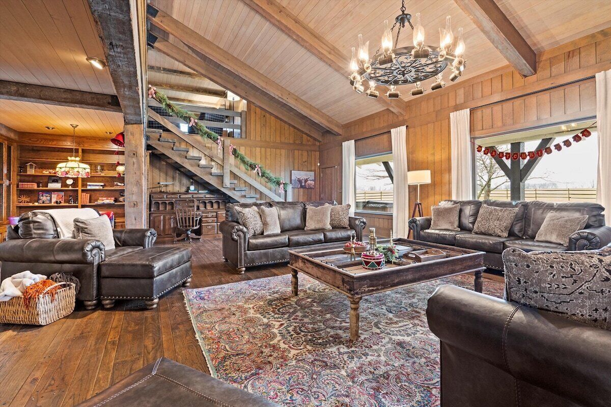 A spacious living room with a grand staircase, leather sofas, a chandelier, and a large window.