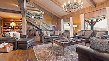 A spacious living room with a grand staircase, leather sofas, a chandelier, and a large window.