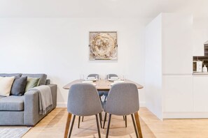 Una sala de estar moderna con un sofá gris, una mesa de comedor con sillas y una cocina con armarios blancos.