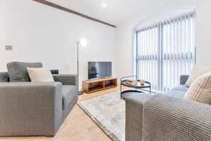 A modern living room with a grey sofa, a flat-screen TV, a floor lamp, and a large window with blinds. - Spacious & Bright 2 Bed Burton-on-Trent Apartment (Burton upon Trent)