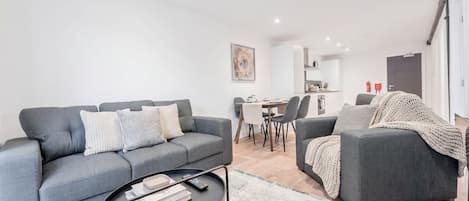 Un salon moderne avec un canapé gris, une table basse et, au fond, un coin repas.
