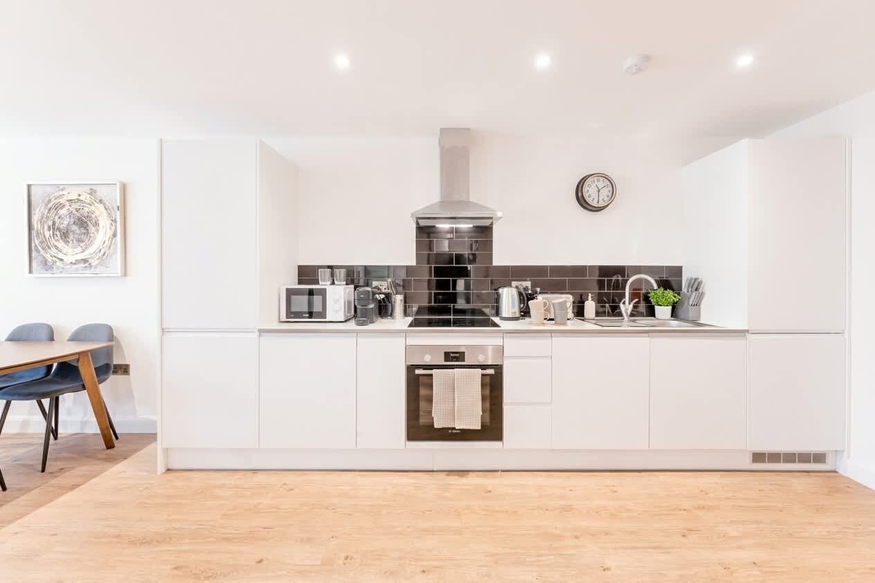 A modern kitchen with white cabinetry, a stainless steel range hood, a built-in oven, and a dining area with a table and chairs.